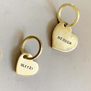 May include: Two heart-shaped brass keychains with attached metal rings. One keychain is stamped with the name BLITZI and the other with NERUDA. A 10 euro cent coin is placed next to the keychains for scale, showing their relative size.