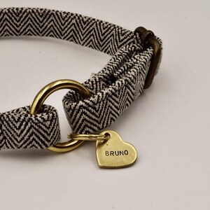 K&ouml;nnte beinhalten: Ein Haustierhalsband mit einem Stoffband in schwarz-wei&szlig;em Zickzackmuster und messingfarbenen Metallbeschl&auml;gen, bestehend aus einem runden Ring und einem herzf&ouml;rmigen Metallanh&auml;nger, der mit dem Namen BRUNO in Gro&szlig;buchstaben graviert ist.