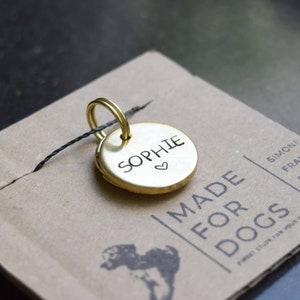 May include: A gold-coloured metal dog tag with the name "Sophie" engraved on it, along with a small heart symbol. The tag is attached to a black string and is sitting on a brown paper background with the text "MADE FOR DOGS" printed on it.