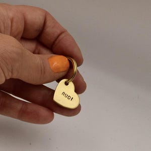 May include: A gold-toned heart-shaped metal keychain tag featuring the name RUDI stamped in black capital letters. The heart charm is attached to a circular metal split ring, providing a functional and simple design for holding keys securely.