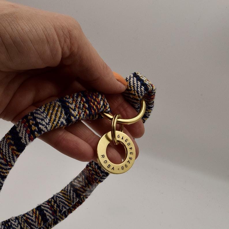 K&ouml;nnte beinhalten: Ein gewebtes Stoffhalsband mit einem mehrfarbigen Zickzackmuster in Marineblau, Wei&szlig; und Orange. Das Halsband verf&uuml;gt &uuml;ber einen polierten goldfarbenen Metallring, an dem eine runde goldfarbene Metallmarke mit der Gravur ROSA und 0676340319 h&auml;ngt.