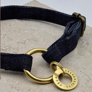 May include: A dark blue denim collar featuring a brass metal ring and a circular brass tag. The tag is engraved with the name ROSA and the number 0676 3403 195. The collar includes a brass slider for adjustment and a sturdy metal D-ring.