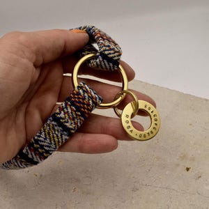 K&ouml;nnte beinhalten: Ein gewebtes Stoffband mit einem mehrfarbigen Muster aus blauen, wei&szlig;en und orangefarbenen F&auml;den, das an einem gro&szlig;en Messingring befestigt ist. Ein kleinerer runder Messinganh&auml;nger ist am Ring befestigt, graviert mit dem Namen ROSA und 08763403195.