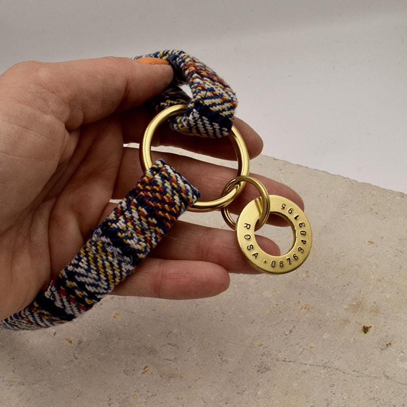 K&ouml;nnte beinhalten: Ein gewebtes Stoffband mit einem mehrfarbigen Muster aus blauen, wei&szlig;en und orangefarbenen F&auml;den, das an einem gro&szlig;en Messingring befestigt ist. Ein kleinerer runder Messinganh&auml;nger ist am Ring befestigt, graviert mit dem Namen ROSA und 08763403195.