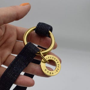 May include: A dark blue fabric strap threaded through a large gold-toned metal ring. A smaller gold-toned metal ring is attached to the larger one, featuring the engraved name ROSA and the numerical sequence 06763403195 along its circular edge.