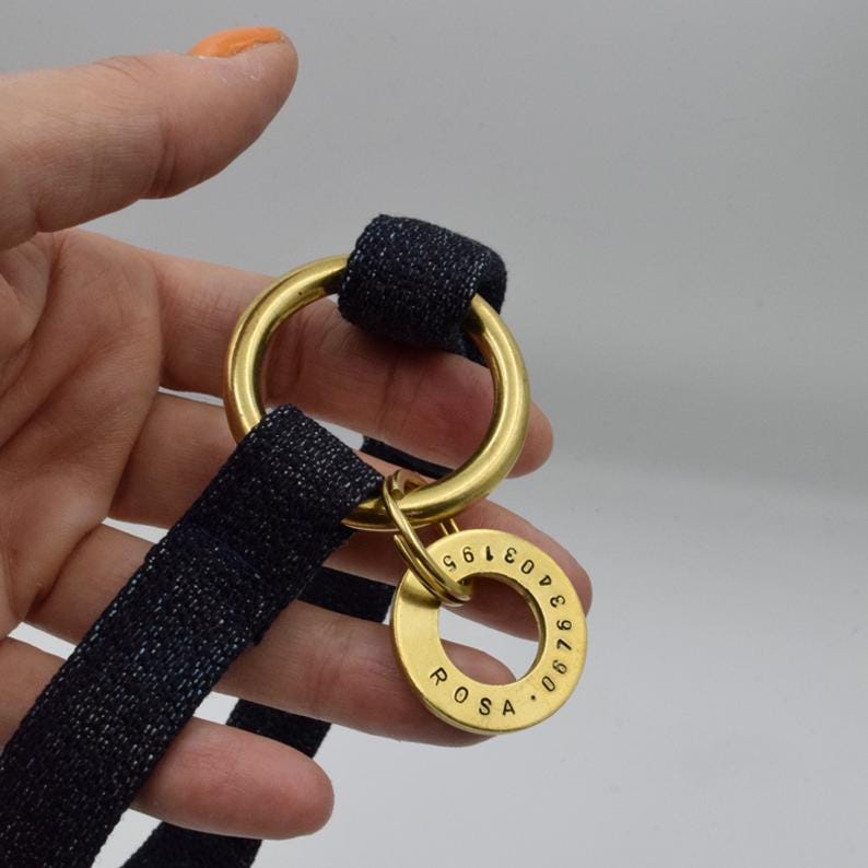 May include: A dark blue fabric strap threaded through a large gold-toned metal ring. A smaller gold-toned metal ring is attached to the larger one, featuring the engraved name ROSA and the numerical sequence 06763403195 along its circular edge.