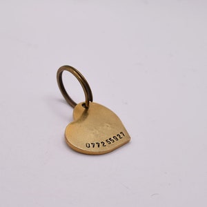 May include: A heart-shaped brass tag with a smooth, matte finish and a circular split ring attached at the top. The tag features the stamped numerical sequence 077255927 along the bottom edge, providing a clear and simple identification marker.