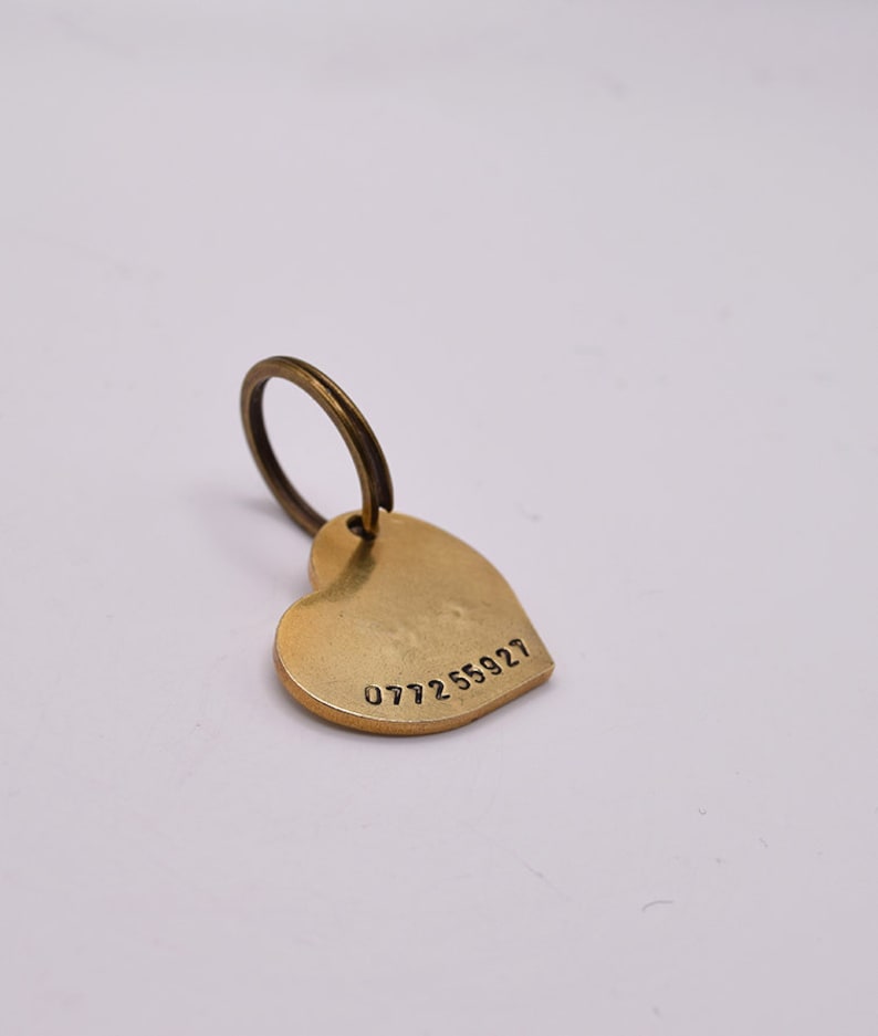 May include: A heart-shaped brass tag with a smooth, matte finish and a circular split ring attached at the top. The tag features the stamped numerical sequence 077255927 along the bottom edge, providing a clear and simple identification marker.
