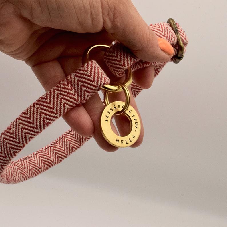 May include: A red and white chevron patterned fabric collar featuring a circular brass tag engraved with the name HELLA and a series of numbers. The collar includes a gold-toned metal D-ring and a matching brass buckle for secure fastening.