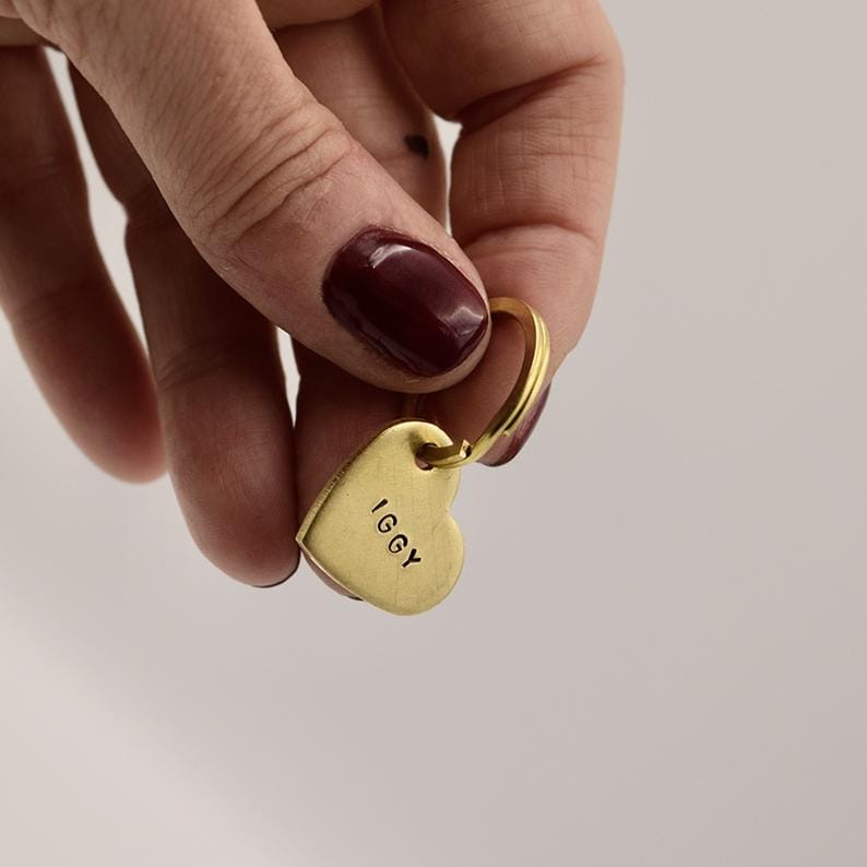 May include: A gold-toned metal heart-shaped charm attached to a circular ring. The heart charm features a smooth, matte finish and has the name IGGY stamped in black capital letters on the front surface. The piece is held by a metal ring.