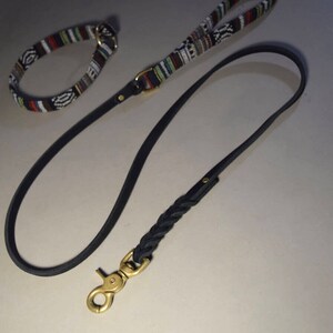 May include: A black leather dog leash with a braided section and a brass clasp. A matching black and white patterned collar is also visible.