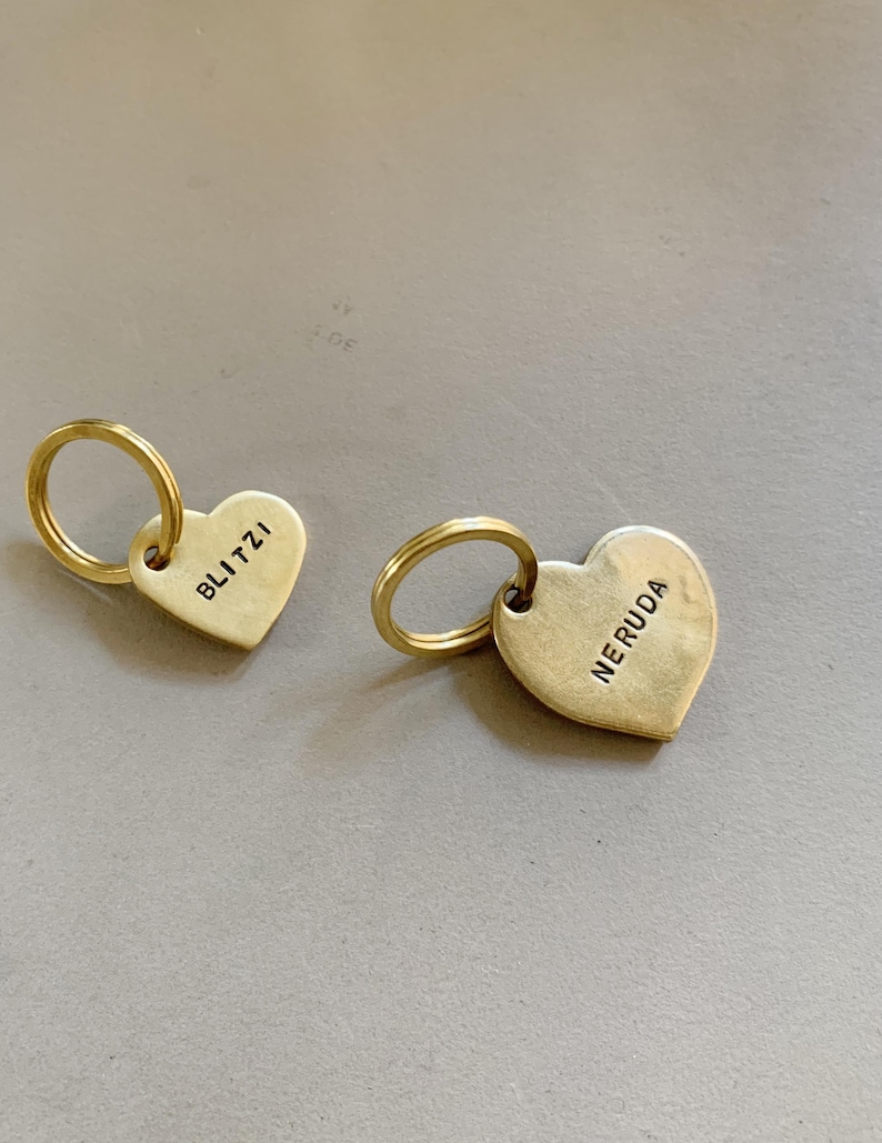 May include: Two gold-toned metal heart-shaped tags, each attached to a circular split ring. One tag is stamped with the name BLITZI and the other tag is stamped with the name NERUDA in black capital letters. The tags have a smooth, matte finish.