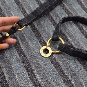 May include: A black textured fabric pet leash and collar set featuring gold-toned metal hardware. A circular gold metal identification tag is attached to the collar ring, engraved with the name ROSA and a series of numbers along the outer edge.