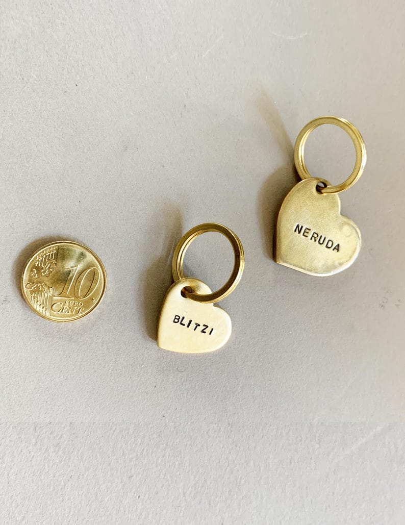 May include: Two heart-shaped brass keychains with attached metal rings. One keychain is stamped with the name BLITZI and the other with NERUDA. A 10 euro cent coin is placed next to the keychains for scale, showing their relative size.