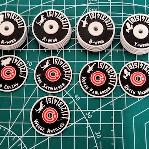 Peut inclure: Ensemble de jetons de jeu noirs et blancs avec des accents rouges et blancs. Chaque jeton présente le nom d'un pilote ou le type de vaisseau, comme "B-WING" et "Keyan Farlander". Les jetons sont disposés sur une grille verte.