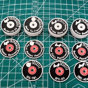 Peut inclure: Ensemble de jetons circulaires noirs et blancs avec des inscriptions variées. Chaque jeton présente un cercle rouge central et du texte blanc, avec des noms tels que "Droid Tri-Fighter" et "Hyena Bomber".