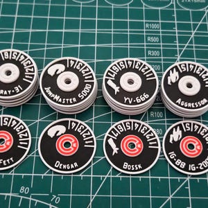 Peut inclure: Huit jetons circulaires noirs et blancs avec divers noms et numéros, dont "Firespray-31", "JumpMaster 5000", "YV-666", "Aggressor", "Boba Fett", "Dengar", "Bossk" et "IG-88 IG-2000". Les jetons sont sur une grille verte.