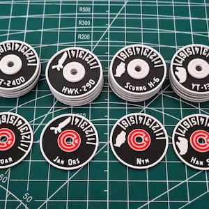 Peut inclure: Huit jetons circulaires noirs et blancs avec divers motifs et textes. Chaque jeton a un anneau extérieur blanc avec des chiffres, un cercle central rouge et une icône blanche. Le texte comprend des noms comme "Han Solo" et "YT-1300".