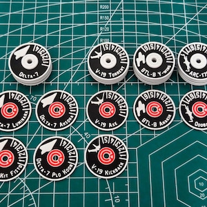 Peut inclure: Une collection de jetons circulaires noirs et blancs avec des accents rouges et blancs. Chaque jeton présente un design et un texte uniques, notamment "DELTA-7", "V-19" et "BTL-B" ainsi que d'autres noms. Les jetons sont disposés sur un tapis de coupe vert.