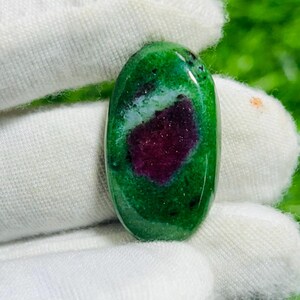 Puede incluir: Una piedra preciosa en forma de cabujón ovalado con una superficie pulida, que presenta un centro de color magenta intenso rodeado de zoisita verde vibrante. La piedra muestra inclusiones oscuras naturales y un acabado liso y brillante.