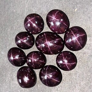 Puede incluir: Un conjunto de gemas pulidas, de forma ovalada y color rojo oscuro. Cada piedra presenta un patrón en forma de estrella con inclusiones de color claro. Las gemas están dispuestas sobre una superficie gris.