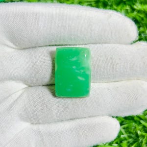May include: A rectangular, bright green gemstone with a slightly translucent appearance. The stone has a polished surface and is held in a white gloved hand. The gemstone has a few white inclusions.