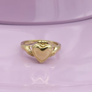 Op de afbeelding: Een goudkleurige ring met een hartvormig medaillon. De ring heeft een gedetailleerd ontwerp met een hartvormig middelpunt en een scharnierende bovenkant. De ring staat tegen een zachte paarse achtergrond, die de ingewikkelde details benadrukt.