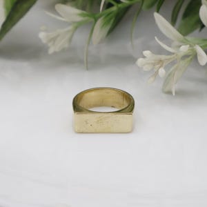 May include: A gold signet ring with a rectangular face. The ring is made of a shiny, metallic material and sits on a white surface. The ring is a simple, classic design.
