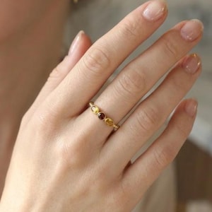 May include: A gold ring with three oval-shaped gemstones in shades of yellow and red. The ring is worn on a finger, showcasing the delicate design and color variations of the stones. The ring is a piece of jewelry.