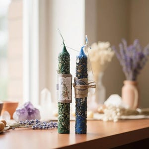 Op de afbeelding: Twee decoratieve pilaar kaarsen, een groene en een blauwe, versierd met gedroogde kruiden en kristallen. De groene kaars heeft een label met botanische illustraties. De kaarsen staan op een houten oppervlak bij een raam, met gedroogde bloemen en kristallen op de achtergrond.