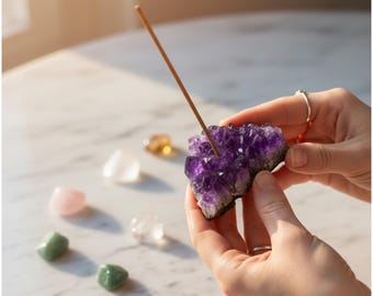 May include: A vibrant purple amethyst crystal cluster with an incense stick inserted. Polished gemstones in pink, green, clear, and yellow are arranged on a white marble surface. The image is bathed in soft, natural light.