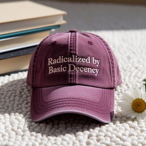 Könnte beinhalten: Burgunderfarbene Baseballkappe mit dem gestickten Text "Radicalized by Basic Decency" in Weiß. Die Kappe liegt auf einer strukturierten Oberfläche, mit Büchern und einer Gänseblümchen im Hintergrund.