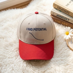 May include: A beige baseball cap with a red peak and a red button. The cap has the navy blue text "I HAD POTENTIAL" and a downward curved line design.