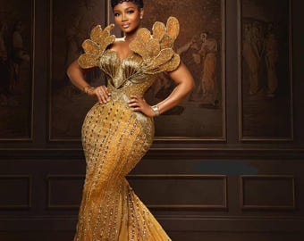 May include: A glamorous, floor-length gold gown with intricate beadwork and a dramatic train. The dress features a plunging neckline and elaborate shoulder embellishments. The woman is posing in front of a dark background with framed artwork.