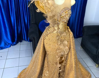 May include: A gold-colored formal gown with intricate floral embellishments and sparkling accents. The dress features a fitted bodice with a sheer overlay and a flowing skirt with a dramatic train. The gown is displayed on a mannequin.