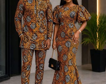 May include: A long-sleeved tunic and matching trousers set alongside a floor-length dress with puff sleeves, both featuring a vibrant orange, black, and white geometric circular pattern. A small black handbag with a gold chain strap is also visible.