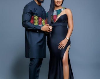 May include: A couple in formal attire. The man wears a dark blue tunic with colorful geometric patterns, paired with matching pants and shoes. The woman is in a black, floor-length gown with a high slit and a patterned shoulder detail.