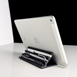 May include: A silver tablet rests on a black tablet stand, with three pens in the stand's slots. The stand is angled, allowing the tablet to be viewed at an angle. The background is black and white.