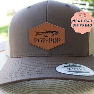May include: A brown and tan snapback hat with a fish graphic and the text "POP-POP" on a leather patch. The hat has a curved brim and a mesh back. The hat also has a "Next Day Shipping" sticker.