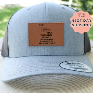 May include: A gray trucker hat with a brown leather patch. The patch features the text "THE Righteous MAN walks in his integrity; his children are blessed after him Proverbs 20:7". The hat has a black mesh back and a curved brim.