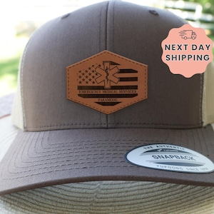 May include: A brown and tan snapback hat with a leather patch featuring an American flag design and the words "Emergency Medical Services Paramedic". The hat has a curved brim and a mesh back.