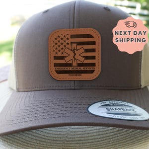 May include: A brown and tan trucker hat with a leather patch featuring an American flag design and the words "Emergency Medical Services Paramedic." The hat has a curved brim and a snapback closure.