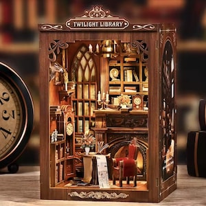 Può includere: Diorama in legno in miniatura della Twilight Library. L'interno presenta scaffali, un camino, una sedia rossa e un tavolo con un giornale. Il diorama include un orologio ed è illuminato da un'illuminazione calda. Il testo "TWILIGHT LIBRARY" è visibile.