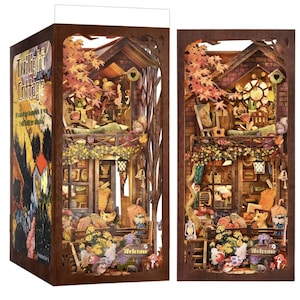 May include: A detailed wooden diorama of a cottage with the text "Twilight Cottage". The cottage features multiple levels, autumn foliage, and intricate cut-out designs. The scene includes miniature furniture, plants, and decorative elements, creating a whimsical, storybook aesthetic.