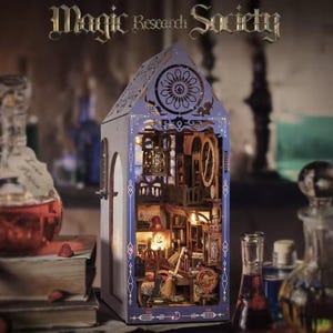 May include: A miniature, illuminated model of a magic research society building. The structure is a light purple with intricate cut-out designs and detailed interior elements. The text "Magic Research Society" is above the building. Various potion bottles are in the foreground.