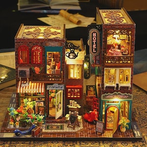 Puede incluir: Una maqueta en miniatura iluminada de una calle de la ciudad con tiendas y un hotel. Los edificios son de ladrillo y madera, con ventanas y toldos detallados. El modelo incluye una floristería, una librería y un hotel con un letrero de neón.