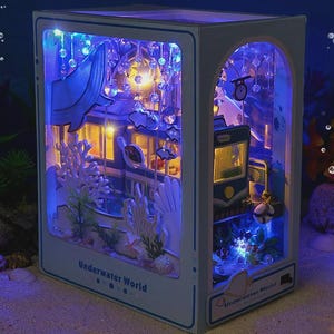 May include: A miniature, illuminated diorama of an underwater scene. The box-shaped display features a detailed depiction of an underwater world with coral, fish, and a building, all lit with blue and yellow lights. The text "Underwater World" is visible on the front.