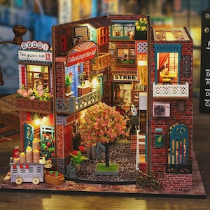 May include: A miniature, detailed diorama of a European street scene. The model features brick buildings, balconies, and street signs, including "GOOD! Time grocery store" and "STREE". A small tree and miniature figures add to the realistic feel. The scene is illuminated with small lights.