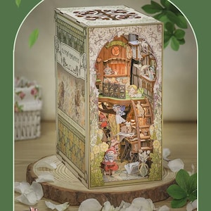 Puede incluir: Diorama de papel detallado, titulado "Elven Paradise", que muestra un mundo élfico en miniatura con detalles intrincados. La escena incluye pequeñas figuras, escaleras y elementos arquitectónicos. El diorama se exhibe sobre una base de madera, con el texto "Elven Paradise".