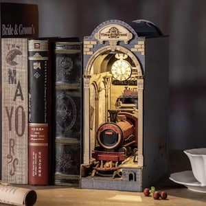 Könnte beinhalten: Ein dekoratives Buch-Eck aus Holz mit einer Bahnhofsszene, einem Uhrturm und einem roten Zug. Die Worte "Time Travel" stehen über der Uhr. Bücher sind links und eine Teetasse rechts.